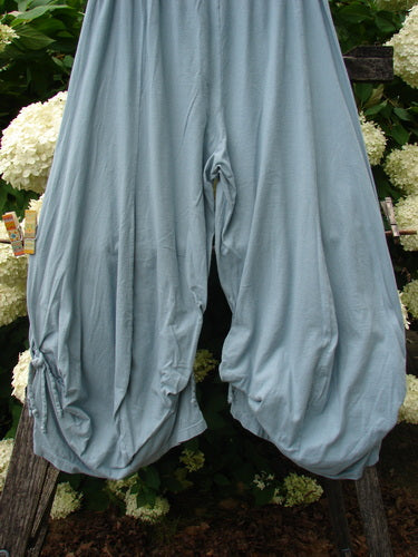 Bluefishfinder.com Barclay Crop Tie Bottom Crop 4 Square Pant in unpainted Blue Cloud, size 0, shown as loose, draped pants with cropped tie-bottom hems. The soft, matte fabric falls in gathered folds, with a pale blue-gray tone set against dense green foliage and white flowers.