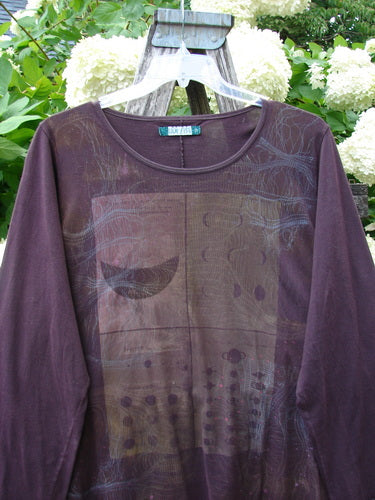 Bluefishfinder.com Barclay Long Sleeved Studio Cafe Tee in Celestial Purple Plum, size 2, with a long-sleeve crewneck silhouette and a muted graphic front in layered plum and brown tones. The soft jersey fabric hangs on a clear hanger against leafy white hydrangeas, creating a natural, garden-bright backdrop.