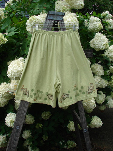 Bluefishfinder.com 1998 Boxer Short Continuous Domino Clover Size 1 in pale olive green, with a wide elastic waistband and loose, airy cut. Small domino and clover motifs trace the lower hem, while the shorts hang from a wooden ladder against white hydrangeas and deep green foliage.