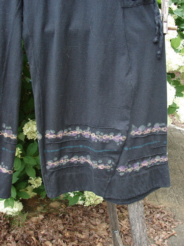 Bluefishfinder.com Barclay Crop Tie Pocket Pinch Pant Wind Chain in black, cropped pants with a relaxed drape and tie waist, finished with subtle floral chain trim near the hem. The dark fabric hangs against leafy greenery, with soft natural light revealing the texture and stitched pocket details.