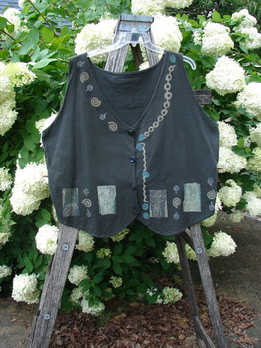 Bluefishfinder.com 1993 The Vest Dancer Black Sand Size 2, a dark sleeveless vest with a deep V neckline and asymmetrical button front. Swirl and circular appliqué details trace the front, while muted patchwork panels line the hem against the weathered wood stand and white flowers.