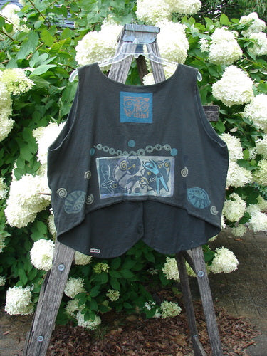 Bluefishfinder.com 1993 The Vest Dancer in black sand, size 2, is a loose sleeveless vest with a soft draped shape and faded blue graphic patches across the front. It hangs on a wooden ladder amid white hydrangeas.