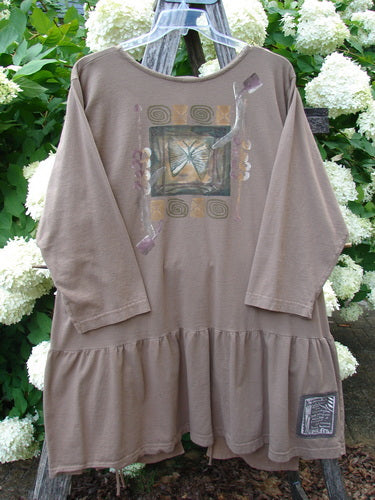 Bluefishfinder.com 1995 Clothespin Jacket Butterfly Mudpie OSFA in taupe, a long-sleeve dress-like garment with a gathered ruffled hem, butterfly graphic, and small patch label. It hangs from a clear hanger before white hydrangeas, with soft natural light and muted earthy tones.
