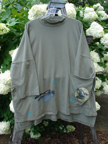 Bluefishfinder.com 1996 Message Top Tumbling Teacup Vine Size 3 in muted olive gray, a loose long-sleeve top with a soft, draped knit and high mock neck. The front shows layered teacup-and-vine graphics near the hem and a large patch pocket, all set against white hydrangeas and green foliage.