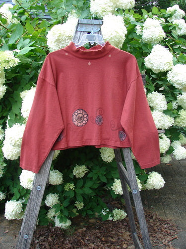 Bluefishfinder.com 1997 Structure Top Continuous Pinwheel Brick OSFA in brick red, a long-sleeve top with a high ruffled collar and subtle circular pinwheel motifs near the hem. It hangs from a wooden ladder amid dense white hydrangea blooms and green foliage, creating a layered outdoor display.