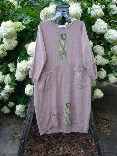 Bluefishfinder.com 1998 Botanicals Thostle Dress Single Tulip Mallow Size 1, a loose mauve linen dress with long sleeves, rounded neckline, and front floral panels. It hangs on a wooden stand before white hydrangeas, its soft texture and muted color palette giving the scene a natural, handmade look.