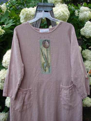 Bluefishfinder.com 1998 Botanicals Thostle Dress Single Tulip Mallow Size 1 in muted mauve, a loose linen dress with long sleeves, gathered side pockets, and a centered botanical tulip print. It hangs on a clear hanger before pale hydrangeas and dark wood, with soft outdoor light.