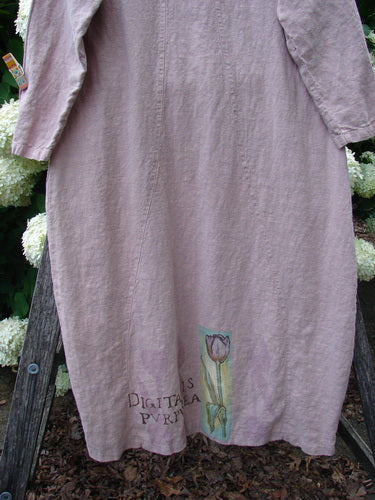 Bluefishfinder.com 1998 Botanicals Thostle Dress Single Tulip Mallow Size 1, a loose lavender-gray dress with long sleeves and soft, wrinkled fabric. A small tulip motif and printed text sit near the hem, framed by dark wooden supports and pale flowers behind it.