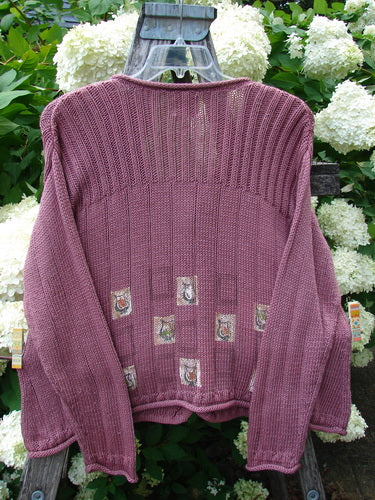 Bluefishfinder.com 1999 Triangle Cardigan Sweater Pitcher Clay Size 1 in dusty mauve knit, with a ribbed high yoke, long sleeves, and small patterned patches across the lower front. It hangs on a dark wooden ladder before dense white hydrangeas and green foliage.