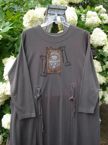 Bluefishfinder.com 1998 Cornucopia Dress Fall Garden Eggplant, size 1, shown as a long-sleeve eggplant dress with a softly draped silhouette and gathered side ties. A centered graphic print adds muted brown and black tones, while the garment hangs outdoors against white hydrangea blooms and green foliage.