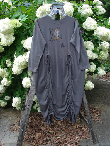 Bluefishfinder.com 1998 Cornucopia Dress Fall Garden Eggplant Size 1 in deep eggplant gray, a loose long-sleeve dress with a round neckline, centered graphic, and gathered draping down the front. It hangs on a wooden easel before white hydrangeas, with soft outdoor light and a muted, earthy palette.
