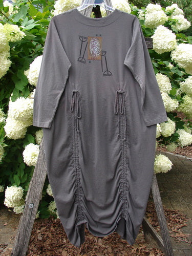 Bluefishfinder.com 1998 Cornucopia Dress Fall Garden Eggplant Size 1 in a muted eggplant-gray tone, made from soft-looking fabric with a loose, long silhouette and long sleeves. A small chest graphic and two gathered drawstring side seams add texture, while the dress hangs from a wooden ladder outdoors against dense white hydrangeas and green foliage.