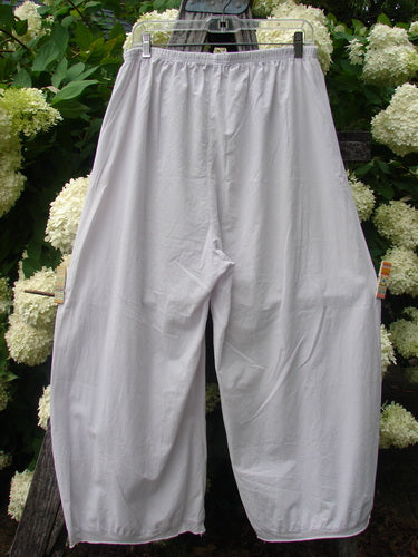 Bluefishfinder.com Barclay Batiste Tiny Flutter Pocketless Pant in unpainted pale pink, shown as loose, wide-leg pants with a soft batiste texture, elastic waistband, and fluttery hem detail. They hang centered against dense green foliage and white blossoms.