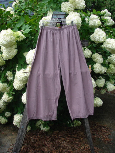 Bluefishfinder.com Barclay Batiste Tiny Flutter Pocketless Pant in unpainted dusty plum, shown as loose, wide-leg pants with a soft batiste texture and flutter hem. The pair hangs from a wooden easel before dense white hydrangeas and green foliage, with muted mauve tones in natural light.