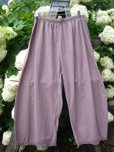 Bluefishfinder.com Barclay Batiste Tiny Flutter Pocketless Pant in unpainted dusty plum, a pair of loose, drapey pants with a gathered elastic waistband and fluttery wide legs. The soft fabric falls from a hanger, with no pockets and a front slit, against leafy white flowers.