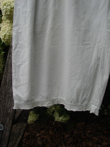 Bluefishfinder.com Barclay Batiste Tiny Flutter Pocketless Pant in unpainted white, shown as a loose, softly draped garment with a straight hem and subtle flutter edging. The lightweight fabric hangs against a rustic backdrop of weathered wood and greenery.