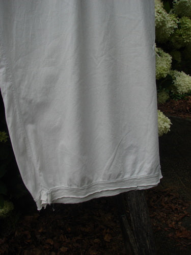 Bluefishfinder.com Barclay Batiste Tiny Flutter Pocketless Pant in unpainted white, shown as a lightweight, softly draped garment with a fluttery hem and pocketless cut. The smooth fabric hangs in a clean vertical panel, with dark foliage behind it adding contrast.