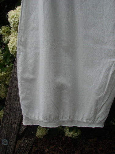 Bluefishfinder.com Barclay Batiste Tiny Flutter Pocketless Pant in unpainted white, size 1, shown in a soft, lightweight fabric with a straight drape and flutter hem. The pant fills the frame against dark wood and pale flowers, creating a muted contrast.