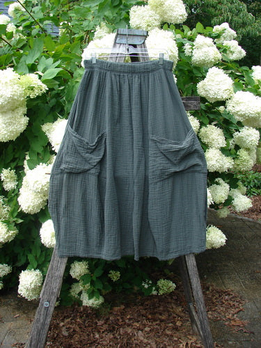Bluefishfinder.com Barclay Gauze Crop Mojo Pant in unpainted evergreen, size 1, shown as loose, cropped gauze pants with a softly crinkled texture, elastic waistband, and gathered patch pockets. The dark green fabric hangs from a wooden stand against white hydrangeas.