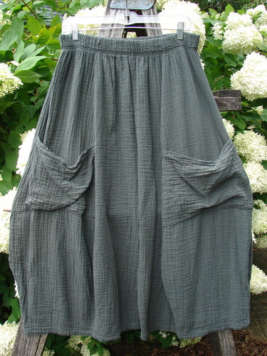 Bluefishfinder.com Barclay Gauze Crop Mojo Pant in unpainted evergreen, size 1, shown as loose, cropped pants with a soft gauze texture, elastic waistband, and layered side pockets. The muted gray-green fabric hangs in gentle folds against a leafy outdoor backdrop.