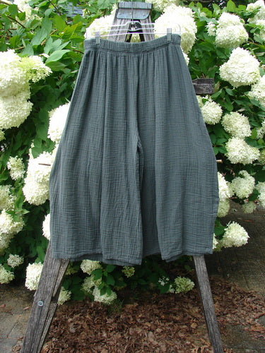 Bluefishfinder.com Barclay Gauze Crop Mojo Pant in unpainted evergreen, size 1, shown as loose, cropped pants in textured gauze with a softly gathered elastic waistband and wide legs. The muted green-gray fabric hangs from a wooden frame against white hydrangeas and dense foliage.