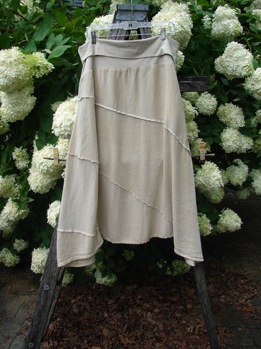 Barclay Hemp Silk Fold Over Diagonal Skirt in unpainted wheat, size 2, with a soft draped silhouette and diagonal seam detail. The lightweight hemp-silk fabric hangs from a wooden hanger, framed by white hydrangea blooms and dark foliage.