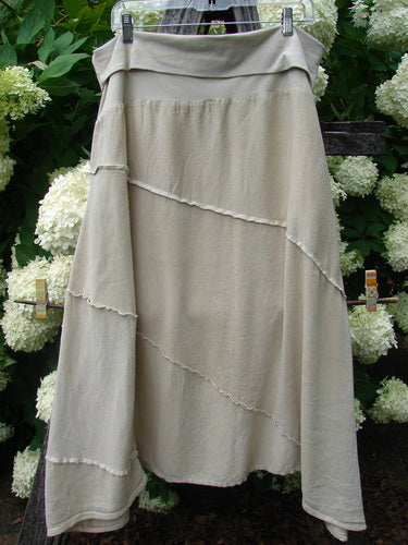 Barclay Hemp Silk Fold Over Diagonal Skirt in unpainted wheat, size 2, with a soft draped fold-over waistband and layered diagonal panels edged in raw, frayed seams. The pale natural fabric hangs in a relaxed A-line shape against dense green foliage and white flowers.