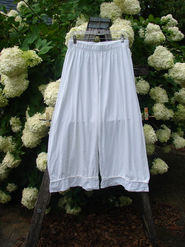 Barclay Flutter Bottom Pocketless Pant in unpainted white, size 1, with a wide-leg silhouette, soft drape, and fluttered hem. The lightweight fabric hangs from a hanger against dense white hydrangeas and dark greenery.