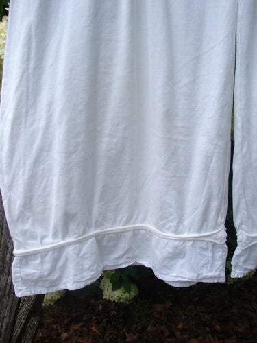 Barclay Flutter Bottom Pocketless Pant in unpainted white, size 1, shown in a soft cotton-like fabric with a fluttered hem and subtle seam detailing. The loose white panel hangs in the foreground, with darker foliage and weathered wood faintly visible behind it.