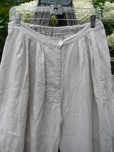 Magnolia Pearl Homespun Linen Ruffle Eyelet Bloomer in natural, OSFA, with a gathered elastic waistband, a single front button, and softly pleated linen fabric in a loose, airy silhouette. The pale neutral cloth hangs from a clear hanger against a blurred floral backdrop.