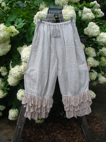 Magnolia Pearl Homespun Linen Ruffle Eyelet Bloomer in natural OSFA, with a loose gathered silhouette, soft linen texture, and layered eyelet ruffles at the hem. The pale fabric hangs from a wooden ladder, framed by white hydrangeas and dark green foliage.
