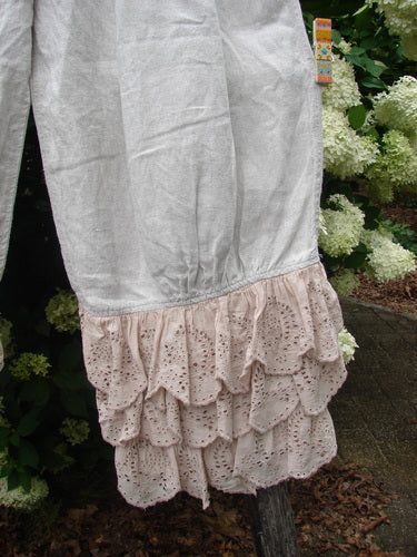 Magnolia Pearl Homespun Linen Ruffle Eyelet Bloomer in natural OSFA, shown with a softly wrinkled linen body and layered eyelet ruffles at the hem. The pale neutral fabric hangs in a loose, airy silhouette against dense green foliage and white blossoms.