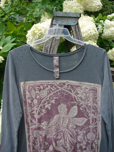 Bluefishfinder.com Magnolia Pearl Cotton Jersey Dylan Dress Fairy Ozzy OSFA in faded charcoal cotton jersey, hanging on a clear plastic hanger with a weathered wooden backdrop. The dress has a round neckline, long sleeves, and a large faded rose-and-fairy graphic in muted mauve and cream.