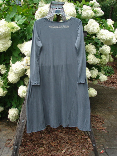 Bluefishfinder.com Magnolia Pearl Cotton Jersey Dylan Dress Fairy Ozzy OSFA in faded blue-gray cotton jersey, with long sleeves, a loose draped silhouette, and Magnolia Pearl lettering at the neckline. It hangs from a wooden stand before white hydrangeas and green foliage, creating a soft outdoor contrast.