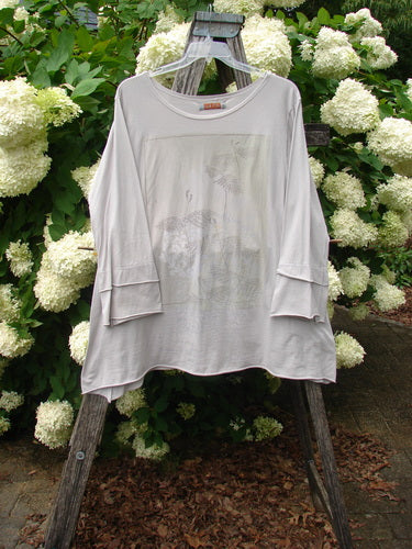 Bluefishfinder.com Barclay Petal Sleeve A Line Tunic Falling Fern Birch Bark Size 1 in soft birch bark, a loose A-line top with wide petal sleeves and a gently curved hem. It hangs on a wooden hanger before pale white hydrangea blooms, with a light, airy print and matte fabric texture.