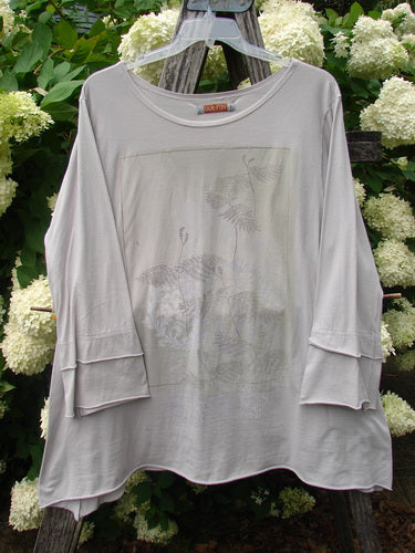 Bluefishfinder.com Barclay Petal Sleeve A Line Tunic Falling Fern Birch Bark Size 1 in soft birch bark gray, with a relaxed A-line silhouette, petal sleeves, and a faint fern print across the front. The lightweight knit drapes loosely over a wooden hanger, set against dense green foliage and cream blossoms.