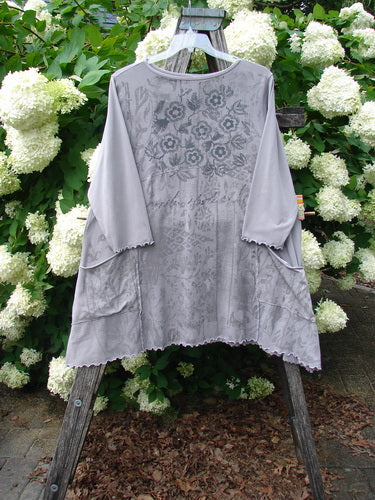 Barclay Cotton Lycra 2 Pocket Urchin Dress Flower Tops Size 0 in light gray, with a loose, flowy silhouette, sheer floral-print yoke, wide three-quarter sleeves, and scalloped edges. The soft cotton-lycra fabric hangs from a wooden hanger against white hydrangeas and green foliage.