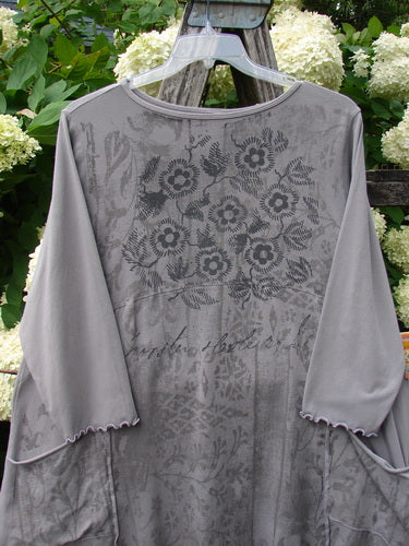 Barclay Cotton Lycra 2 Pocket Urchin Dress Flower Tops Size 0 in soft gray cotton-lycra, with a loose drape, long sleeves, scalloped edges, and faded floral and script prints. It hangs on a clear hanger against leafy greenery.