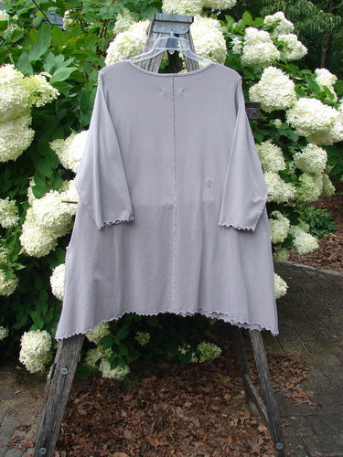 Barclay Cotton Lycra 2 Pocket Urchin Dress Flower Tops size 0 in soft gray, a loose A-line garment with three-quarter sleeves, scalloped hems, and two subtle front pockets. It hangs on a clear hanger outdoors, framed by dense white hydrangea blooms and dark greenery.