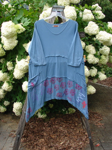 Bluefishfinder.com Barclay Raw Seam Vector Tunic Dress Daisy Folly Echo Blue Size 1, a loose blue tunic dress with raw seam detailing, three-quarter sleeves, and an uneven hem. The soft, layered fabric shows purple floral accents near the lower panel, hanging from a wooden stand against white hydrangea blooms and green foliage.