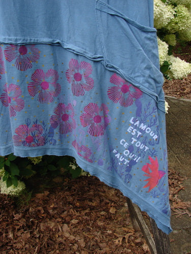 Bluefishfinder.com Barclay Raw Seam Vector Tunic Dress Daisy Folly Echo Blue, a blue layered tunic dress with raw seam edges and a draped asymmetrical hem. The fabric shows magenta daisy-like prints and white graphic text, photographed outdoors against greenery.