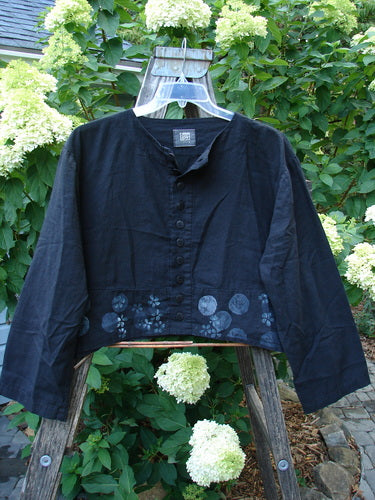 Black 2000 Light Hemp Crop Curve Jacket Moonflower with front variegated buttons, A-line flair, and downward back yoke, hanging on a swinger amidst garden flowers.