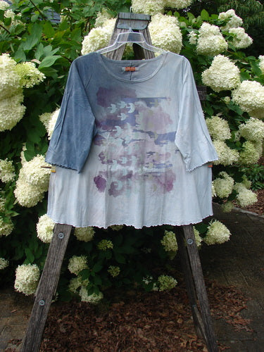 Bluefishfinder.com Barclay Three Quarter Sleeved Cafe Tee in Dove Flight Mottled Grey Blue Cloud, a loose cotton tee with three-quarter sleeves, a faded cloudlike print, and a softly frayed hem. It hangs centered on a wooden stand against dense white hydrangeas and green foliage.