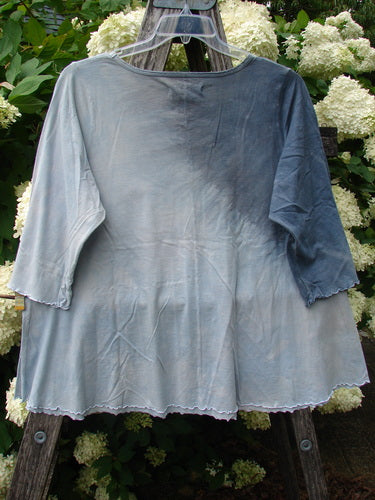 Bluefishfinder.com Barclay Three Quarter Sleeved Cafe Tee in Dove Flight mottled grey blue cloud, size 1, hangs from a clear hanger with a softly draped, three-quarter-sleeve silhouette. The lightweight fabric shows a washed, textured finish and a gently flared hem against pale floral blooms and dark wooden supports.