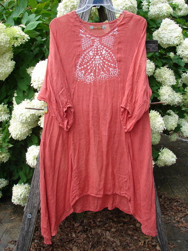 Bluefishfinder.com Barclay Linen High Vent Venetian Dress Center Starburst Poppy Size 1 in faded coral-red linen, with a loose drape, rounded neckline, and white starburst motif centered on the chest. The dress hangs on a wooden stand against dense white hydrangeas and green foliage.