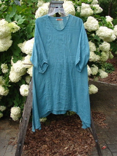 Bluefishfinder.com Barclay Linen High Vent Venetian Dress in unpainted jade, a loose linen garment with a V-neck, elbow-length sleeves, and a high vent hem. It hangs on a wooden stand against white hydrangeas, the textured blue-green fabric reading softly in natural light.