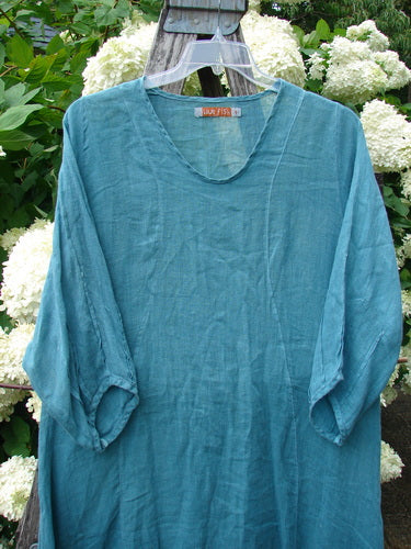 Bluefishfinder.com Barclay Linen High Vent Venetian Dress, unpainted jade, size 1, hangs on a clear hanger with a softly draped linen texture, three-quarter sleeves, and a loose V-neck silhouette. The blue-green fabric fills the frame against white hydrangeas and deep green foliage.