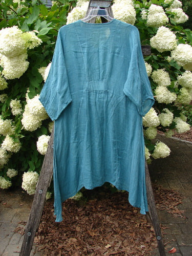 Bluefishfinder.com Barclay Linen High Vent Venetian Dress in unpainted jade, size 1, shown as a loose, airy linen garment with a softly crinkled texture, wide sleeves, and an open high-vent hem. It hangs from a wooden frame before white hydrangeas and green foliage, with muted daylight emphasizing the blue-green fabric.