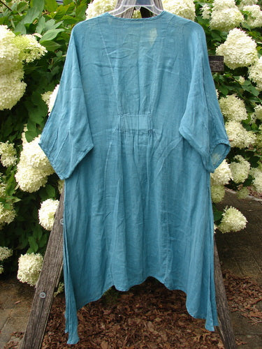Bluefishfinder.com Barclay Linen High Vent Venetian Dress in unpainted jade, size 1, shown as a loose linen garment with a round neckline, wide elbow-length sleeves, and a high vent hem. The airy, textured fabric hangs in soft folds against a leafy outdoor backdrop.