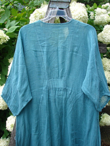 Bluefishfinder.com Barclay Linen High Vent Venetian Dress in unpainted jade, Size 1, hangs on a clear hanger with a softly wrinkled linen texture, wide three-quarter sleeves, and a loose, airy drape. The teal-blue fabric is framed by white hydrangeas and greenery, giving the garment a natural, outdoor presentation.